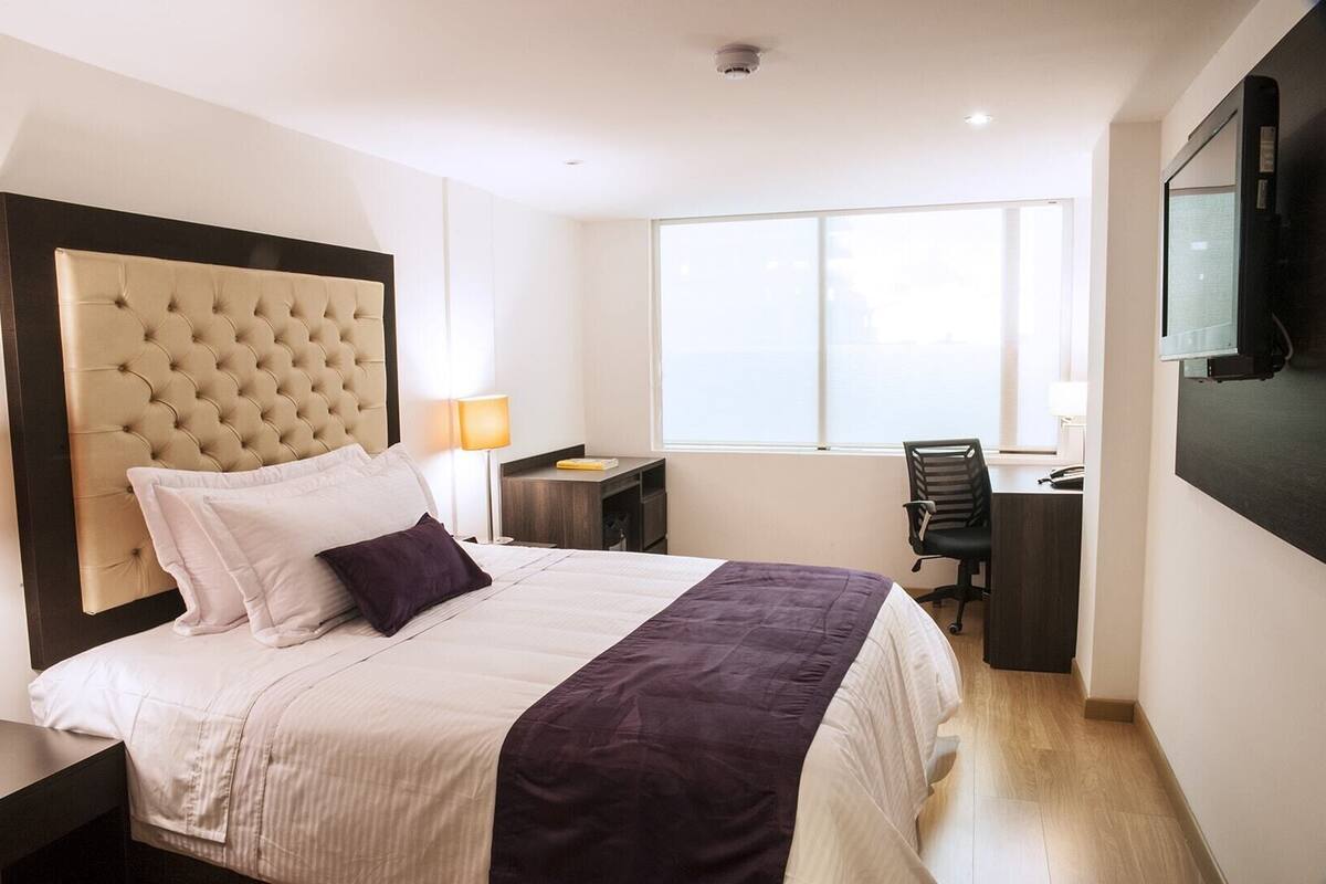 A hotel room with a large bed, a desk, a chair, a TV, and a window.