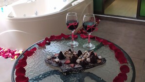 A round table with rose petals and chocolate treats, a bathtub, and a bed with a tufted headboard.
