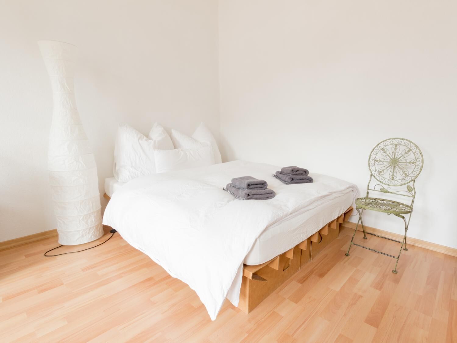 Ein schlicht eingerichtetes Schlafzimmer mit einem hölzernen Bettgestell, weißer Bettwäsche und einem grünen Metallstuhl.