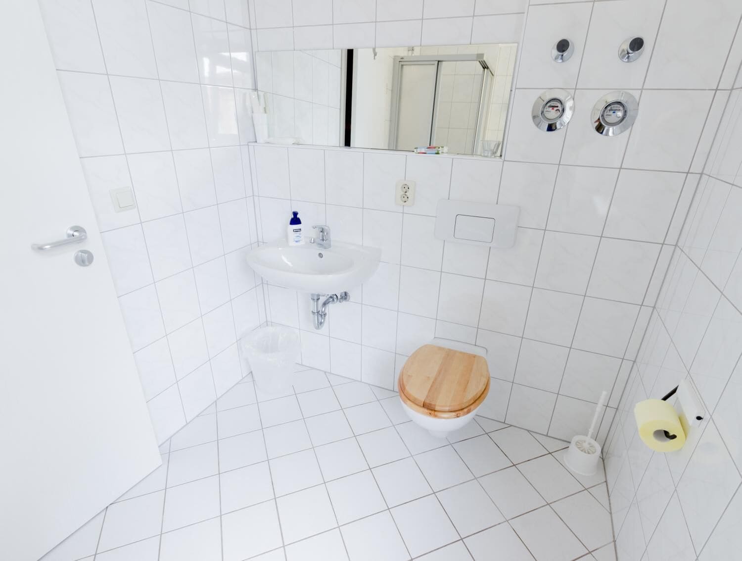 A bathroom with white tiles, a sink, a toilet, a mirror, and a shower with a control panel.