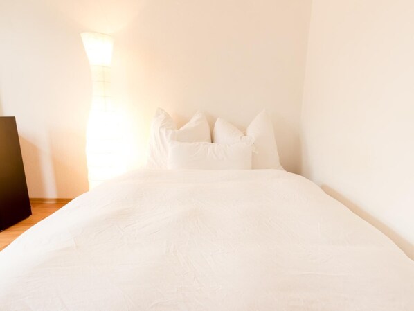 A neatly made bed with white linens and pillows, a bedside lamp, and a wooden nightstand.