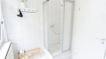 Une salle de bain moderne avec des carreaux blancs, une douche en verre, un banc en bois sur lequel sont posés des articles de toilette et une plante en pot sur une étagère.