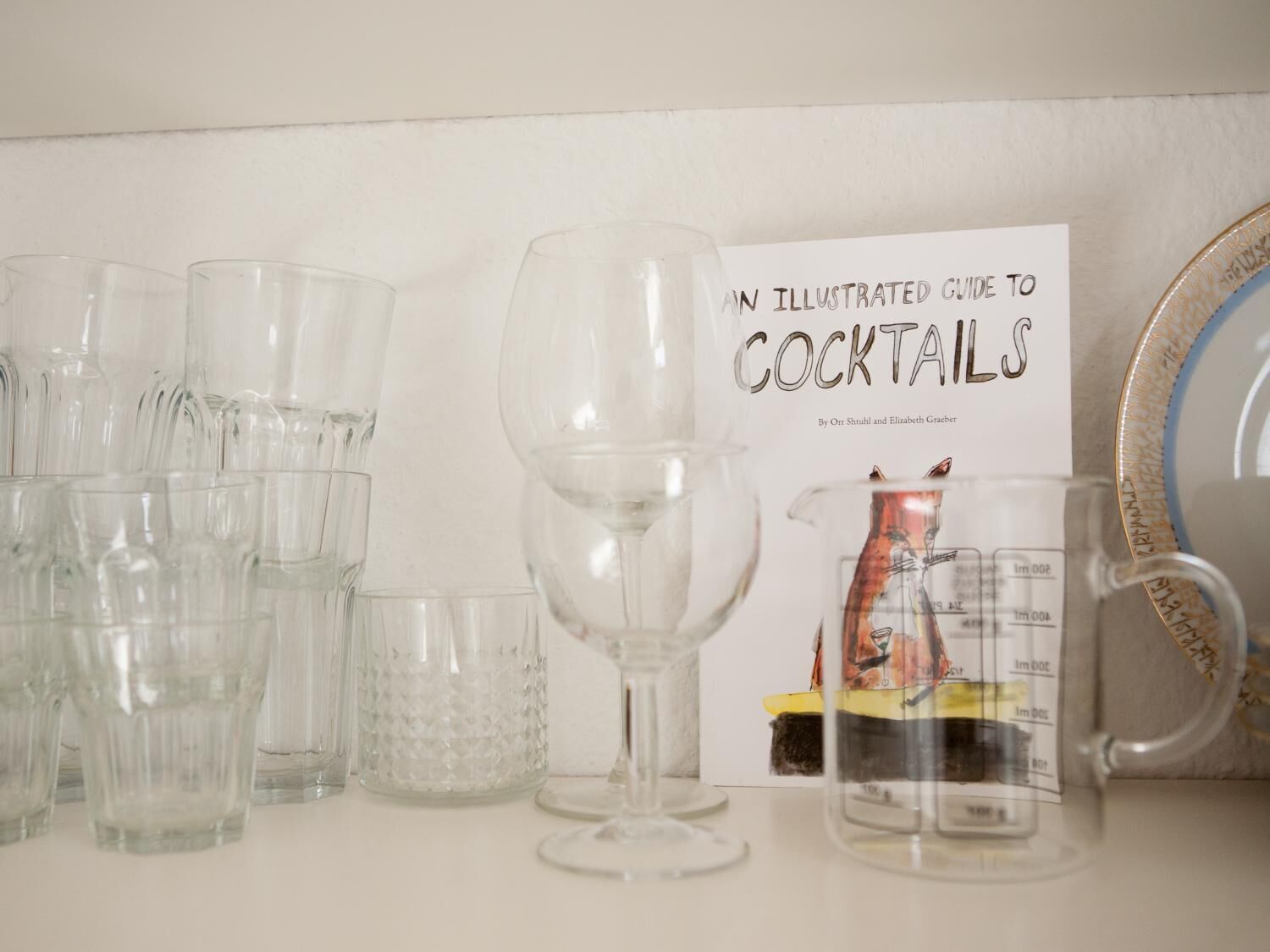 A collection of clear glassware and a book titled 'An Illustrated Guide to Cocktails' by Orr Shuhil and Elizabeth Graebt.