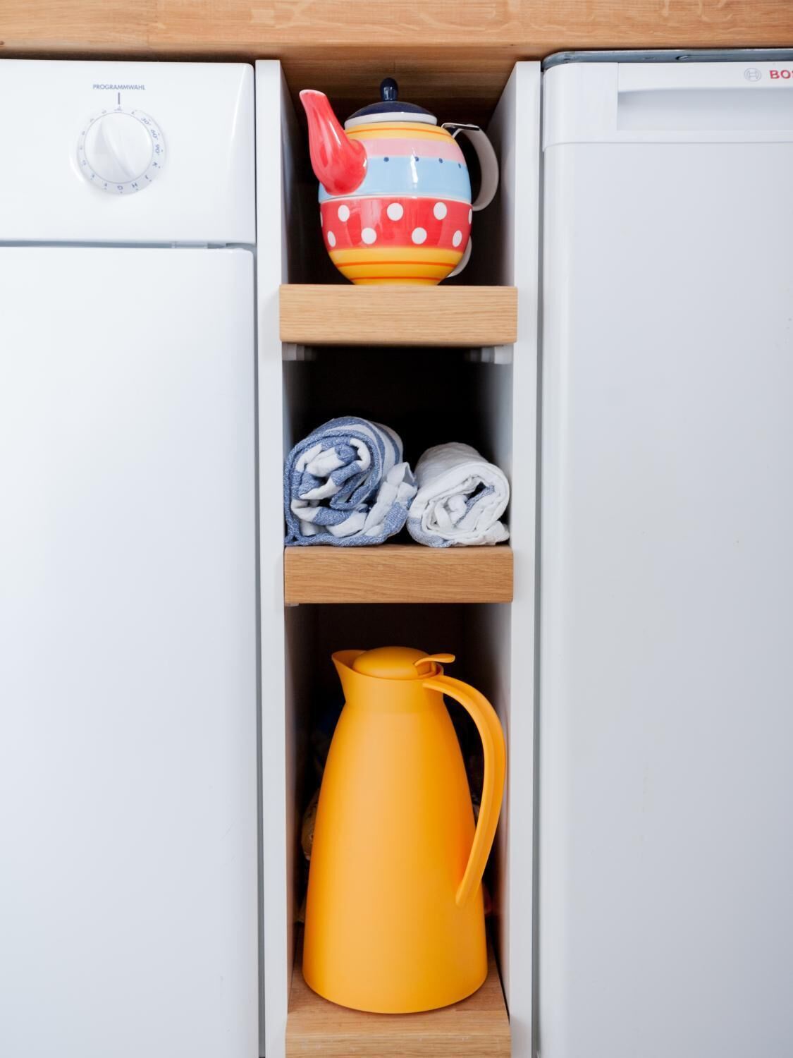 A kitchen area with a white refrigerator, a colorful teapot on a shelf, rolled towels, and a yellow coffee pot.