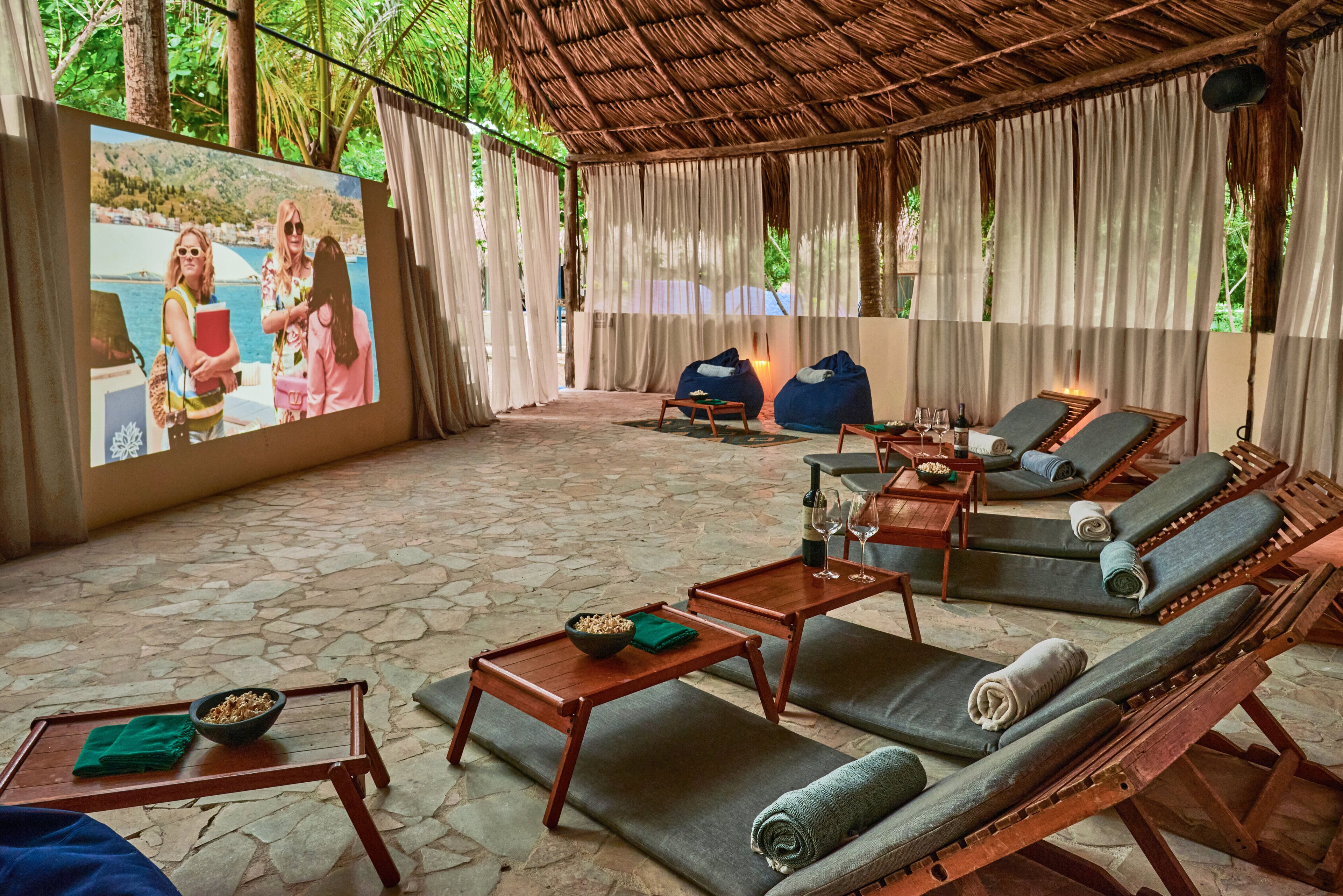 A covered outdoor seating area with stone flooring, wooden furniture, and a large screen displaying a photo.