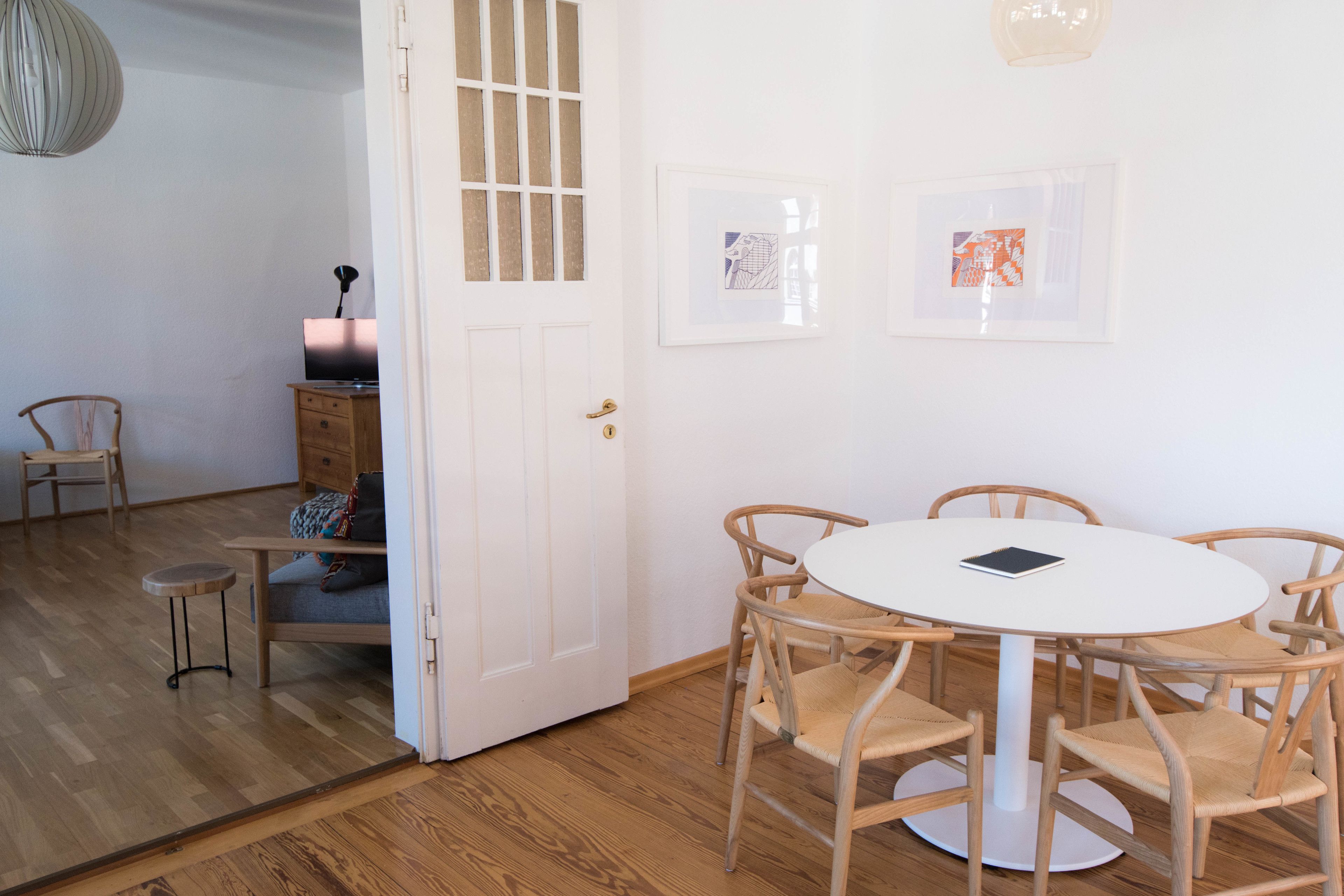 A room with a round table, wooden chairs, a small stool, a door, and two framed pictures on the wall.