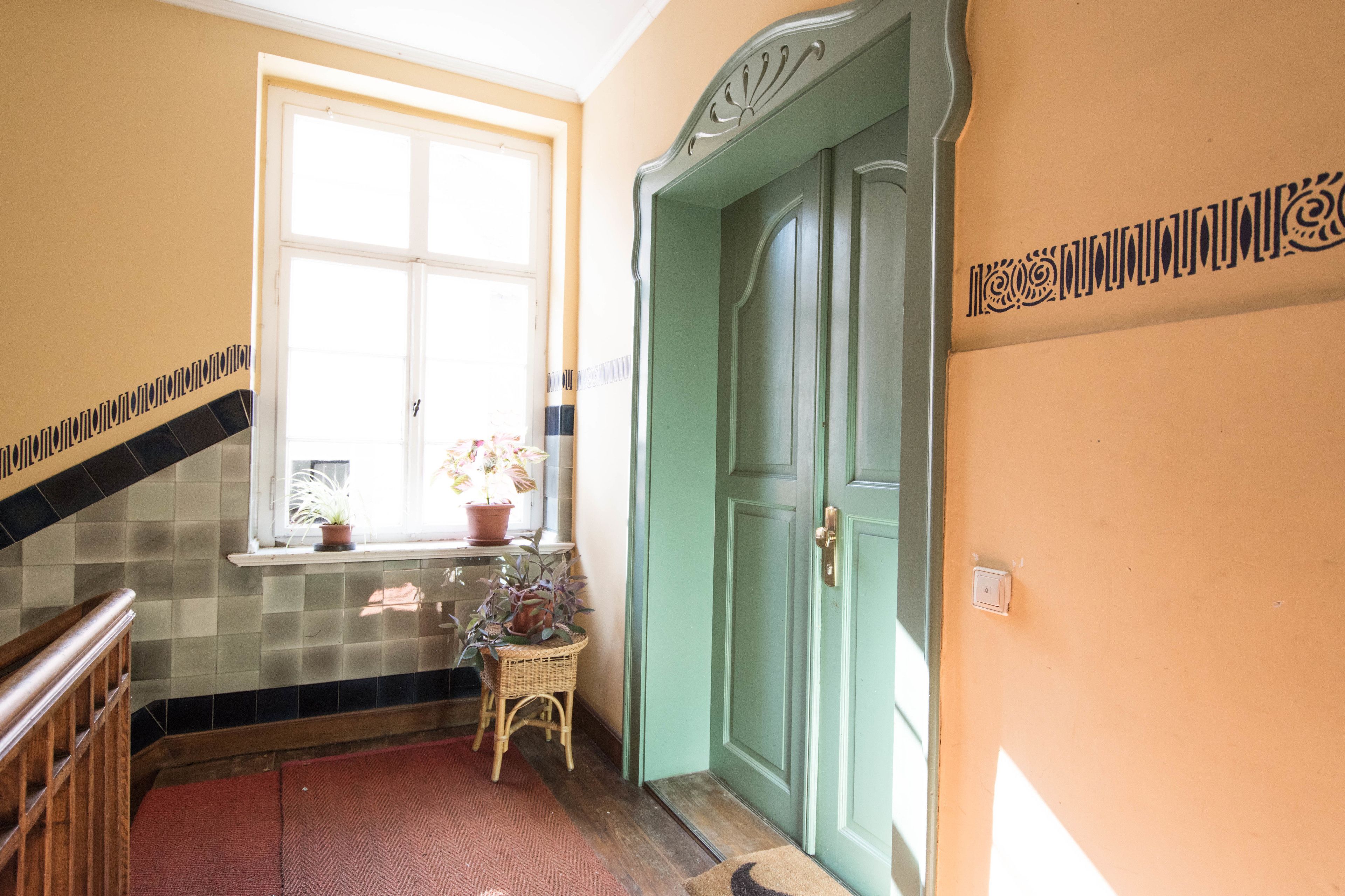 Ein Korridor mit Fliesenboden, einer Holzhandläufe, einem Fenster mit Blumentopf und einer grünen Tür.