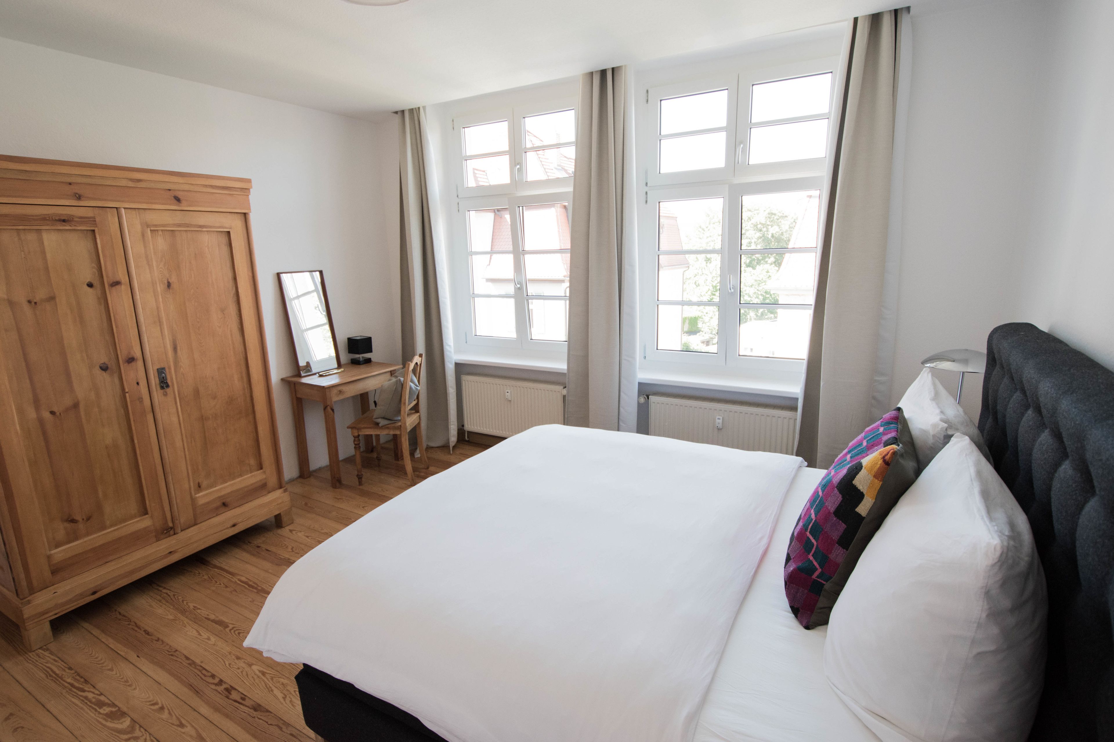 Een slaapkamer met een groot bed, een houten garderobe, een bureau met stoel, en een raam met gordijnen.