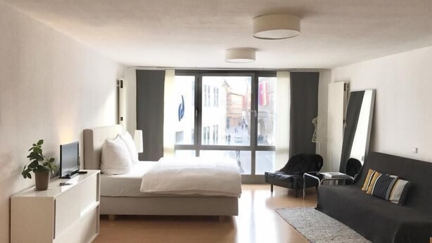 A modern hotel room with a bed, a sofa, a desk with a lamp, and a large window offering a view of the street. - Central Studio Marktplatz (Weimar)