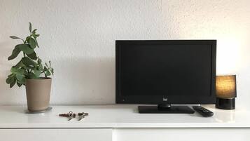 Un escritorio blanco con un monitor negro, una planta en maceta, un control remoto y una lámpara.