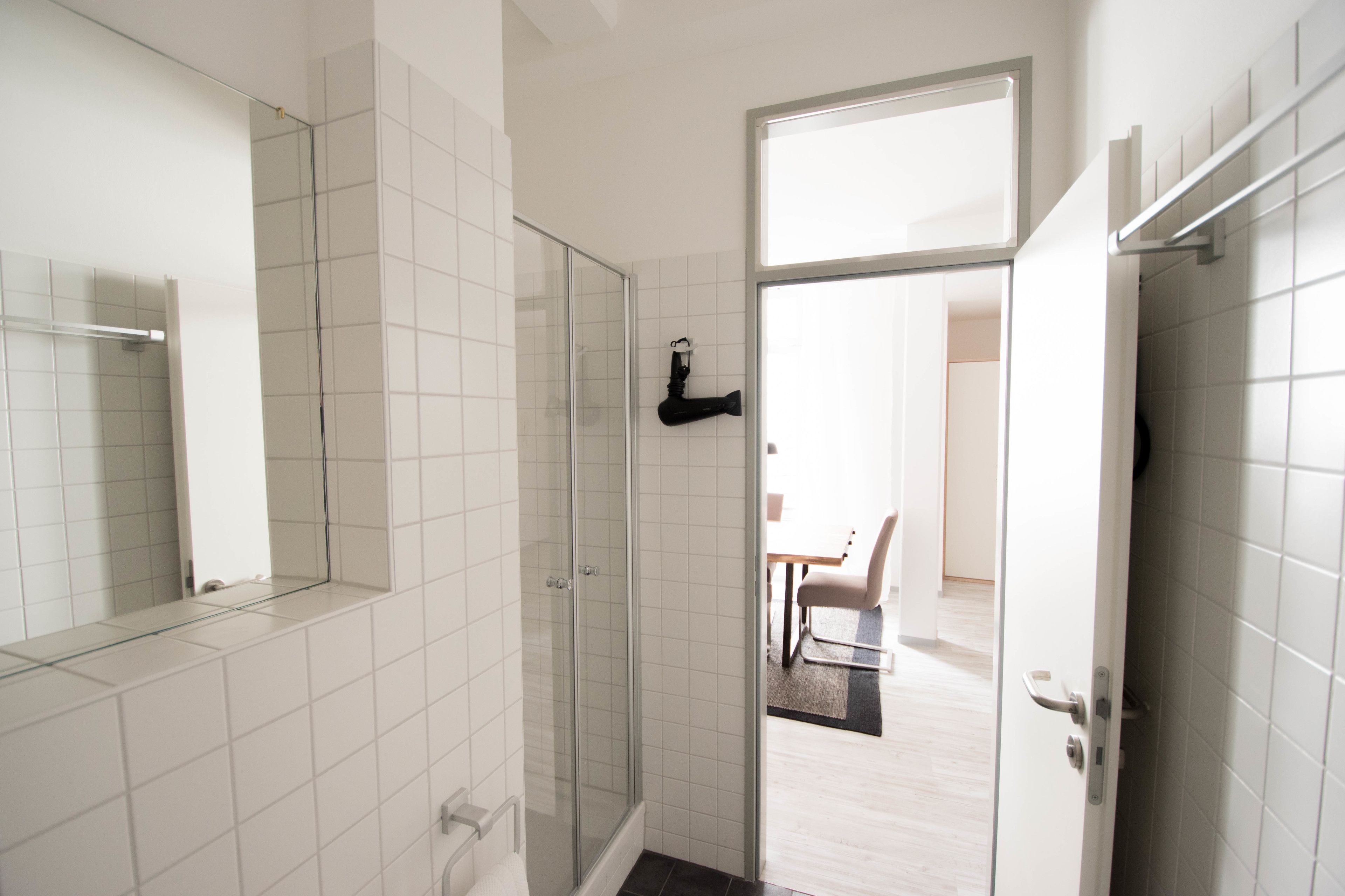 Ein modernes Badezimmer mit weißen Fliesen, einer Duschkabine aus Glas und einer sichtbaren Tür zu einem Essbereich.