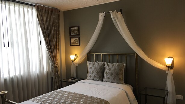 A hotel room with a bed, two bedside tables, a nightstand, a canopy bed, and a window with curtains. - Hostal Mitad del Mundo (Quito)