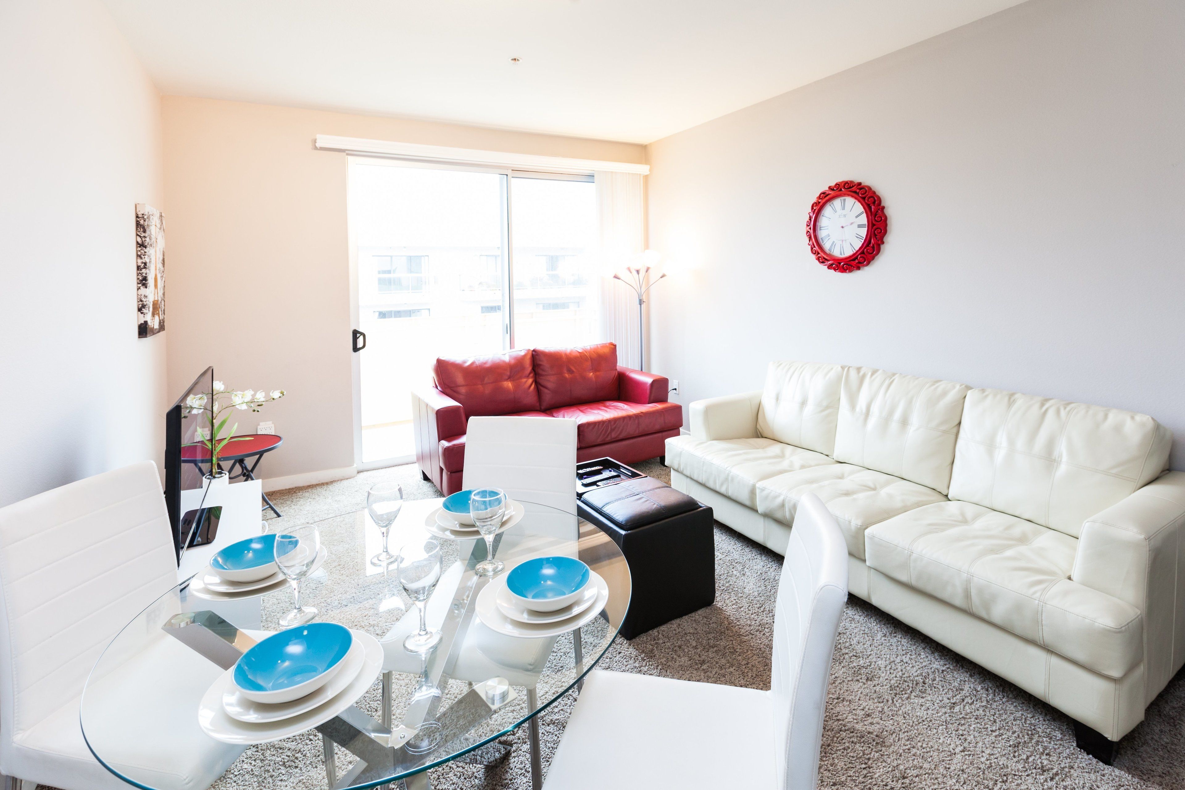 A modern living room with a glass dining table set for four, a red sofa, a white sofa, and a red wall clock.