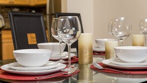 Mesa de comedor lista con cuencos, platos y copas de vino blancas, junto a velas y un mantel individual rojo.