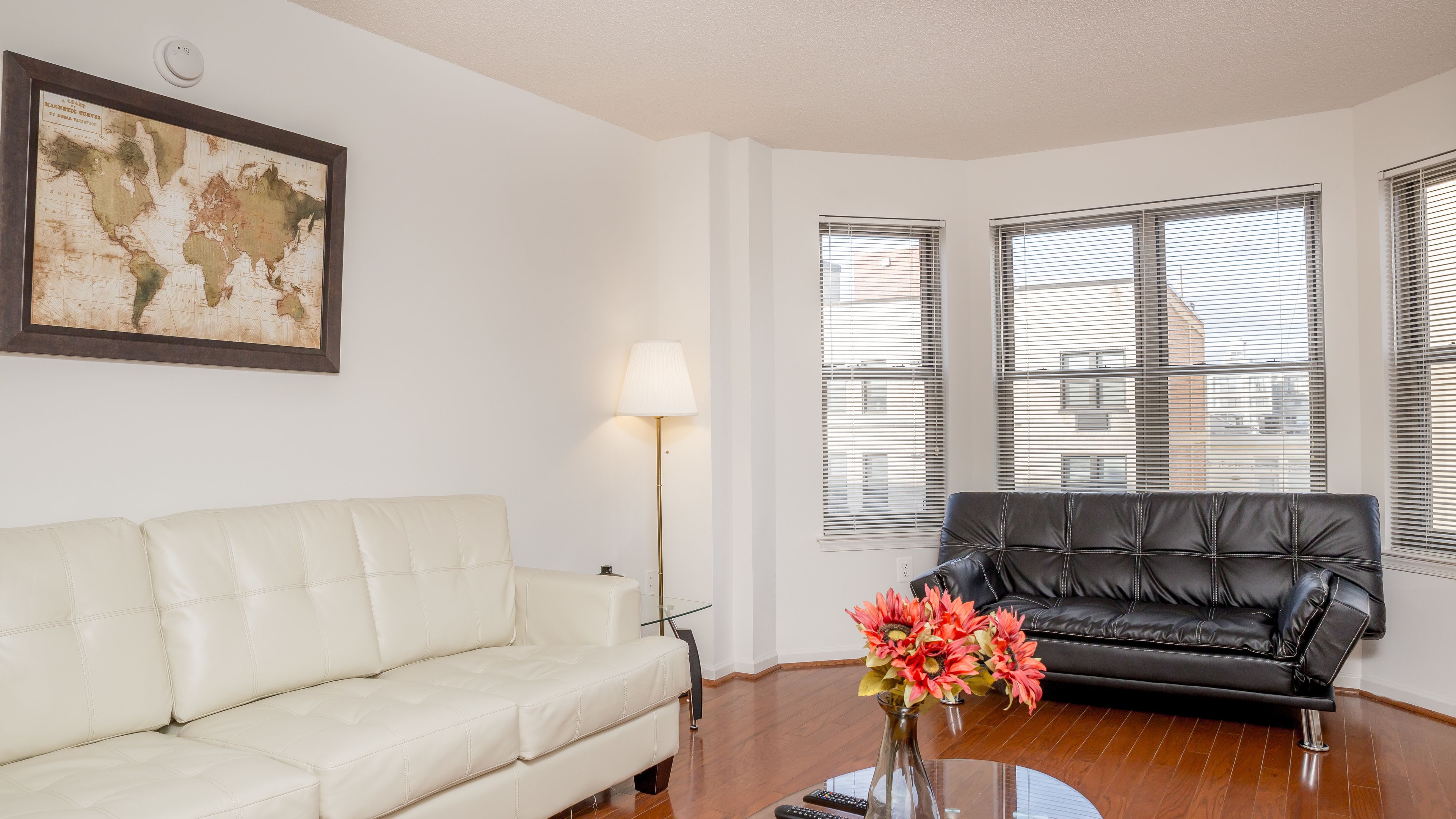 A living room with a white sofa, a black leather sofa, a floor lamp, a vase with flowers, and a framed map on the wall.