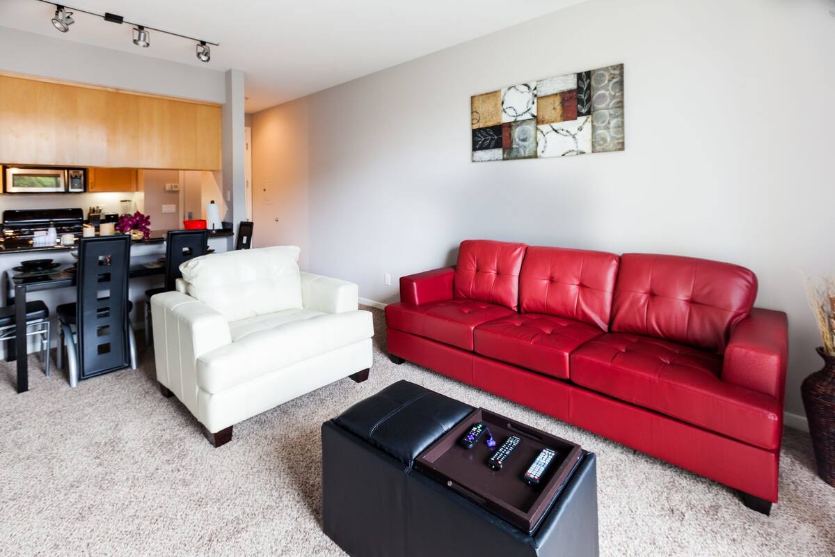 A modern living room with a red sofa, a white armchair, and a black ottoman with remote controls.