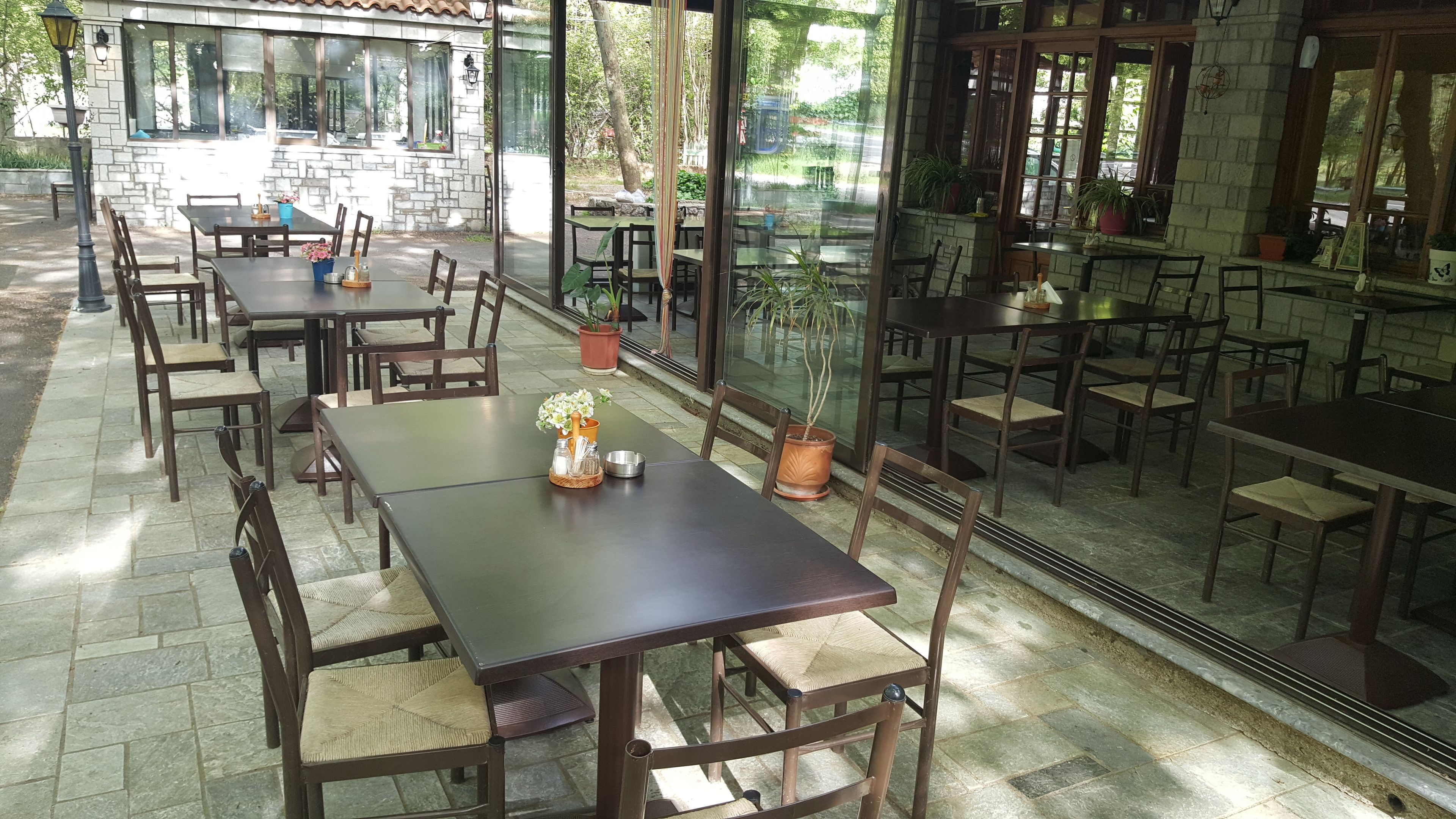 An outdoor cafe with multiple tables and chairs, potted plants, and a stone wall.