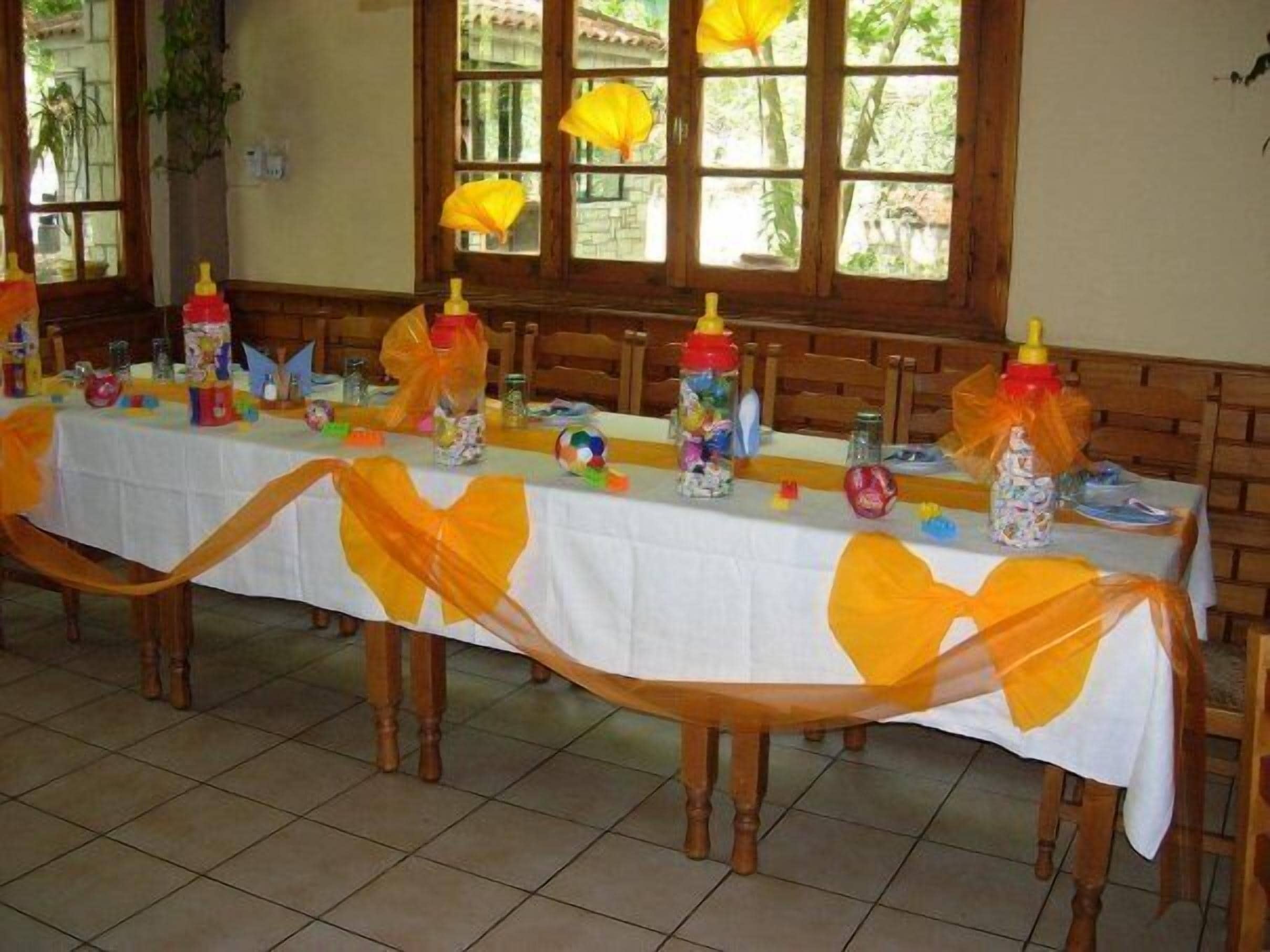 A long dining table with a white tablecloth, decorated with yellow ribbons and various items like bottles and toys.