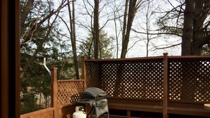 A wooden deck with a grill and a propane tank, surrounded by a lattice fence and trees. - Inn Towne Cottage (Bancroft)