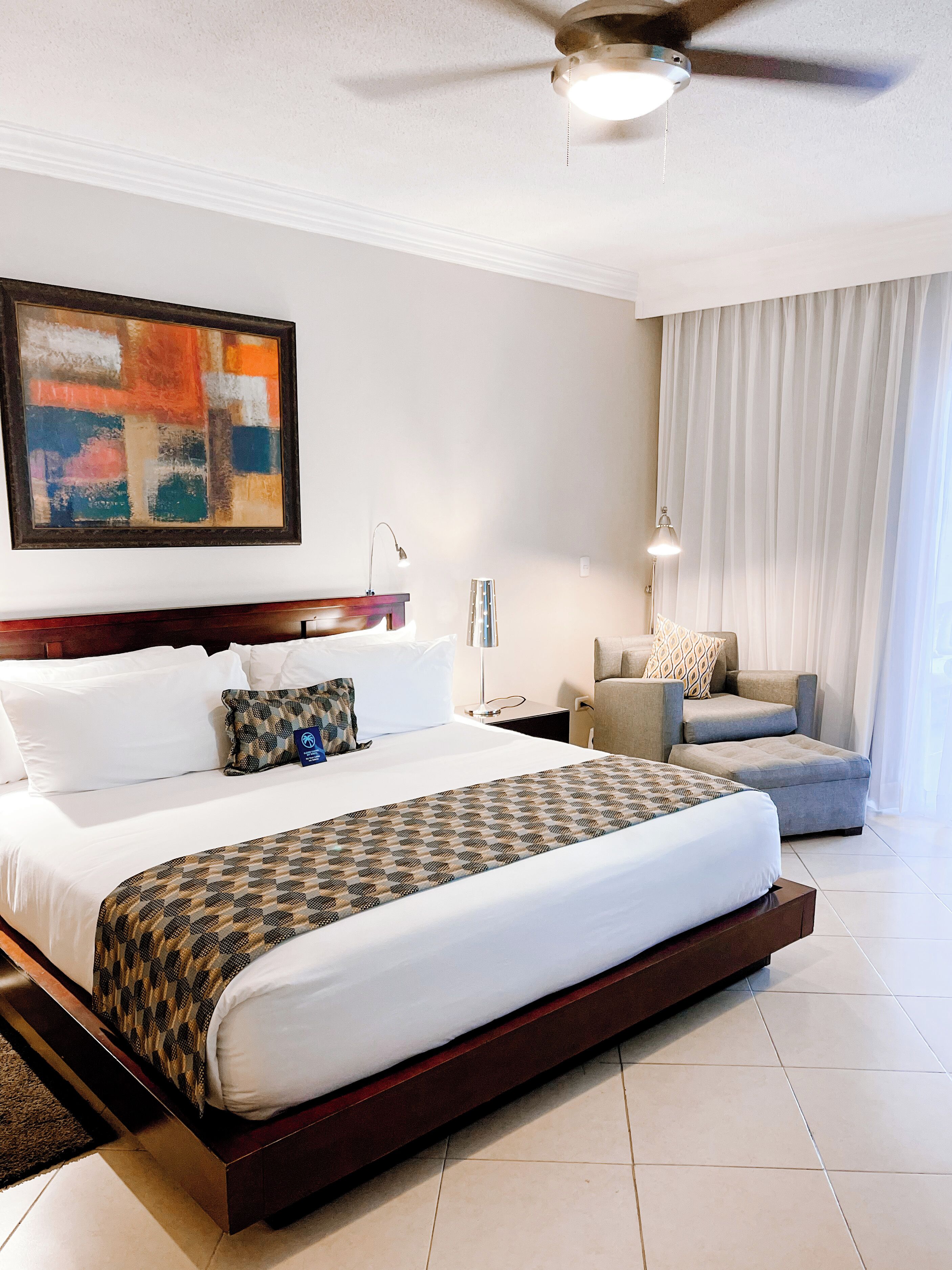 A hotel room with a large bed, a ceiling fan, a painting on the wall, a desk with a lamp, and a view of the outside through the window.