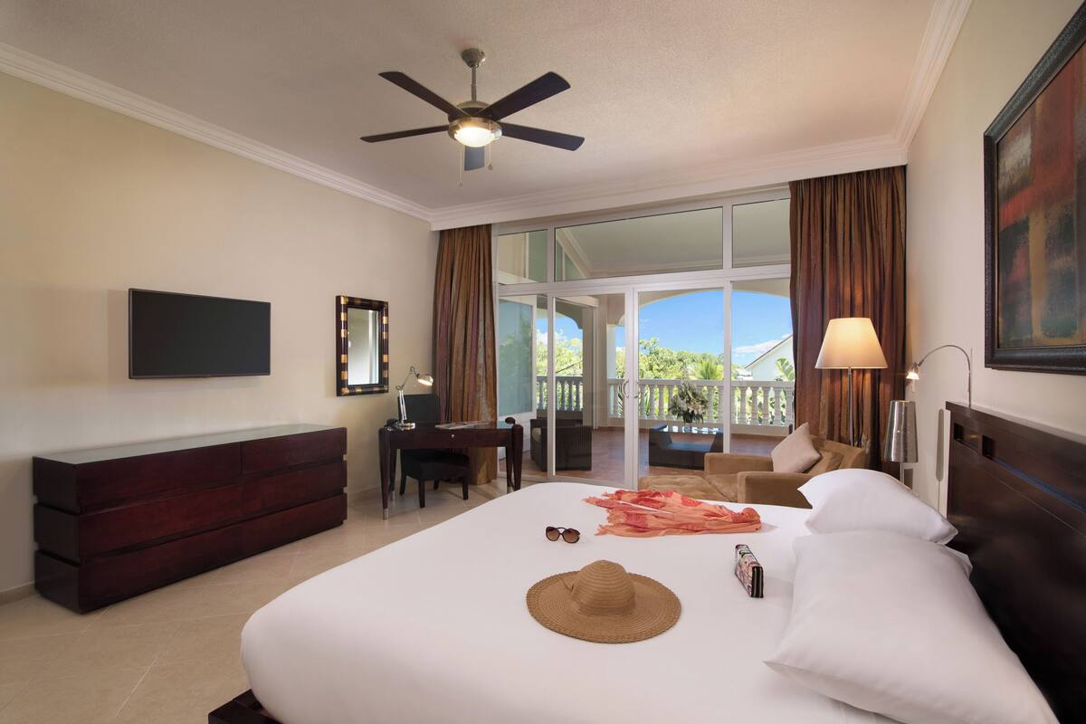 Habitación de hotel con cama, escritorio, silla, televisión y balcón con cortinas.