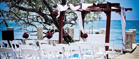 An outdoor wedding ceremony setup with white chairs and a wooden arch, overlooking the sea.