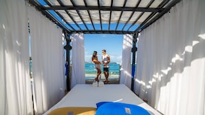 A room with a bed, white curtains, and a view of the beach.