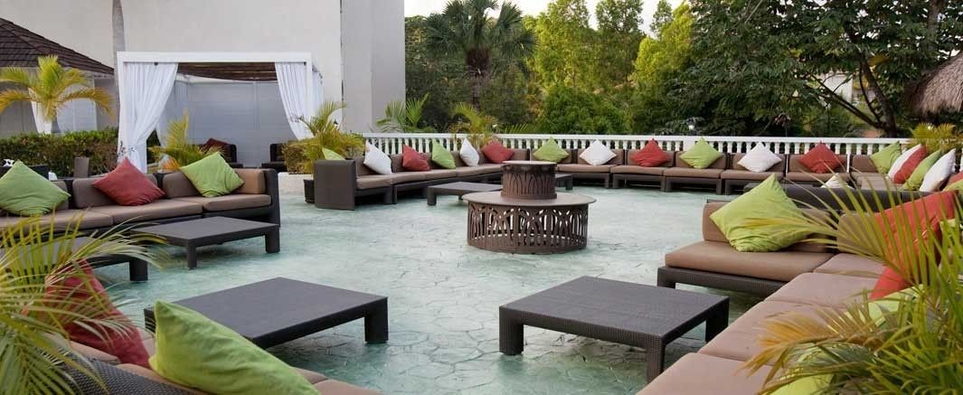 Un espacio al aire libre con sofás y mesas, rodeado de vegetación y una piscina.