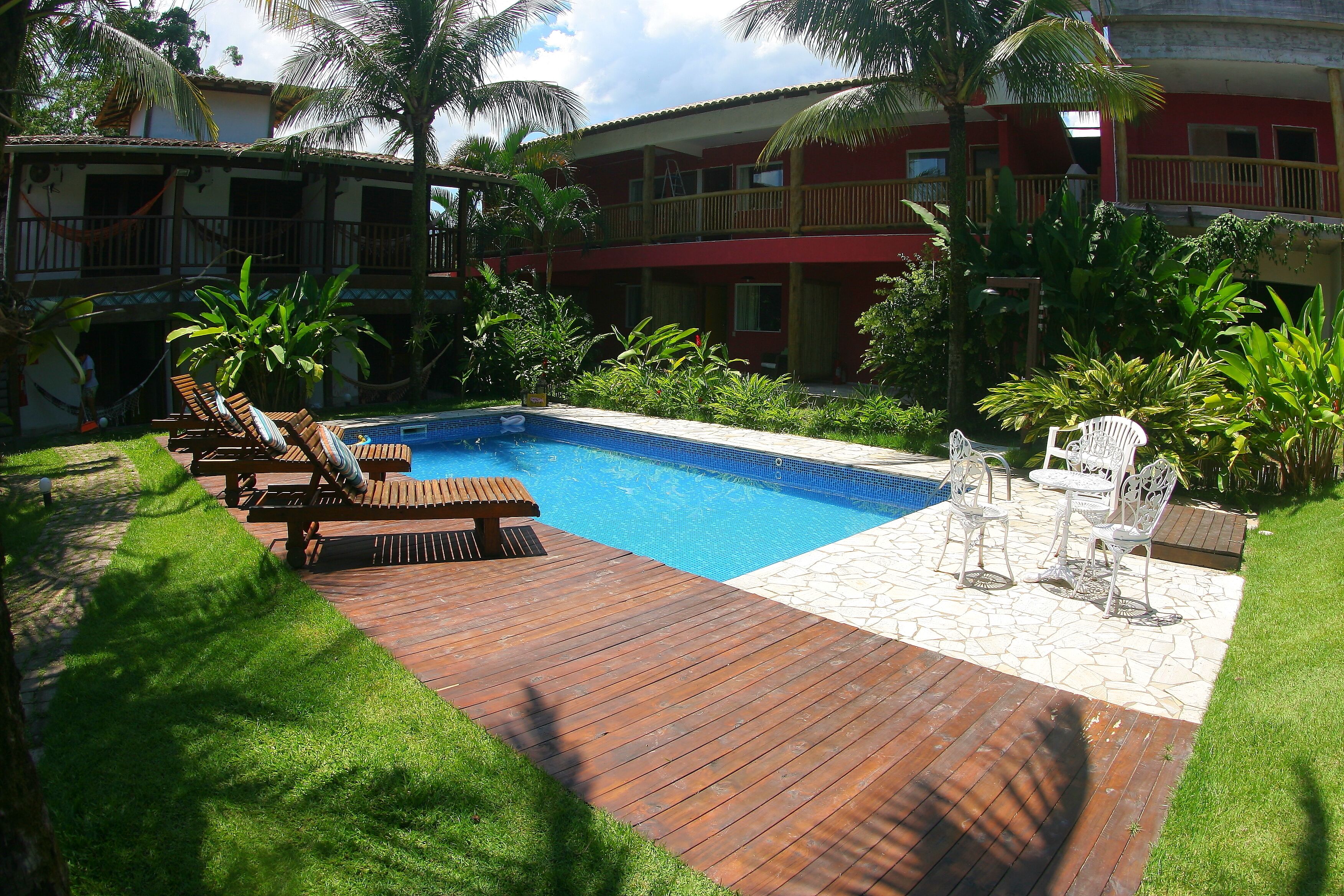 A swimming pool with wooden deck and lounge chairs, surrounded by lush greenery and a red building with balconies.