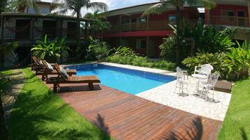 A swimming pool with wooden deck and lounge chairs, surrounded by lush greenery and a red building with balconies.