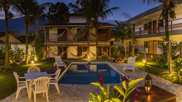 A pool area with lounge chairs and a hammock, surrounded by palm trees and a building with a balcony. - Pousada Tubes - Hostel (São Sebastião)