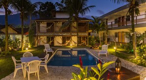 A pool area with lounge chairs and a hammock, surrounded by palm trees and a building with a balcony. - Pousada Tubes - Hostel (São Sebastião)