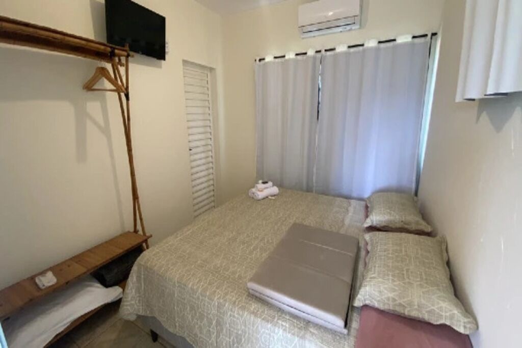 Quarto com cama, prateleira de madeira, televisão na parede e janela com cortinas.