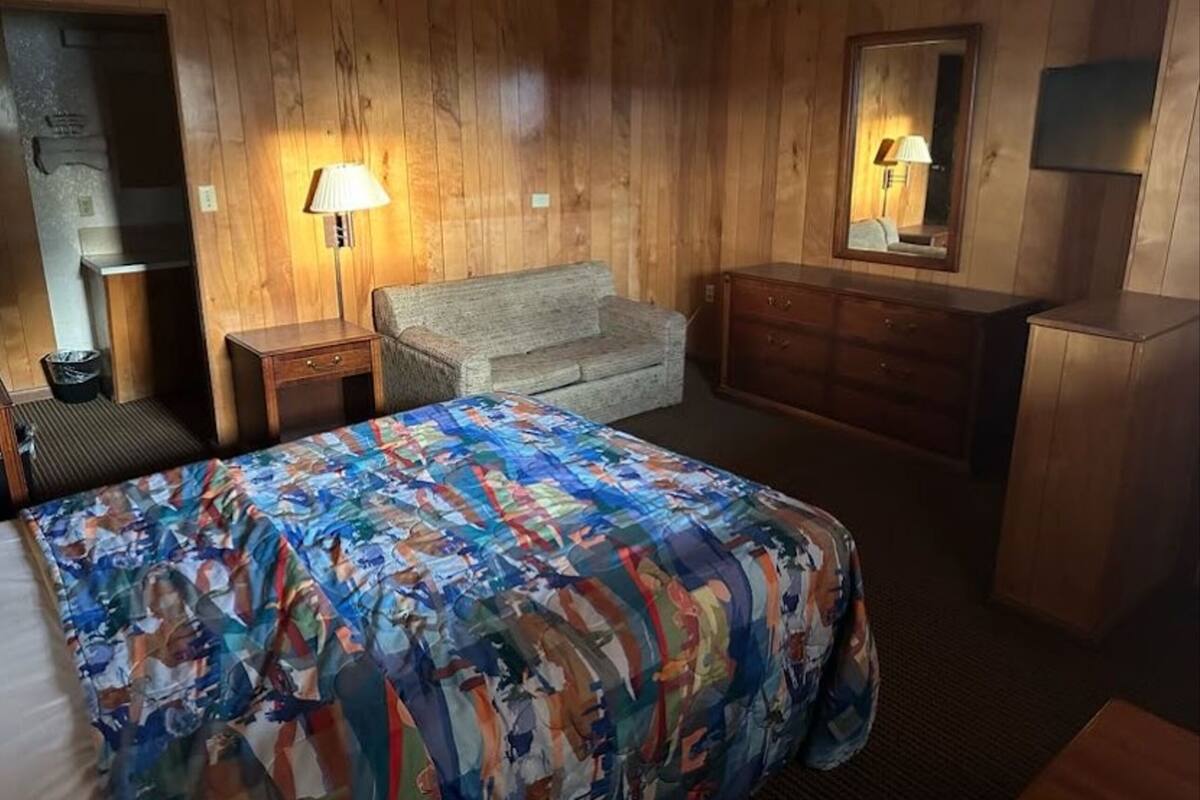A room with wooden walls, a bed with a colorful bedspread, a sofa, a dresser, and a lamp.