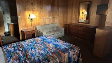 A room with wooden walls, a bed with a colorful bedspread, a sofa, a dresser, and a lamp.