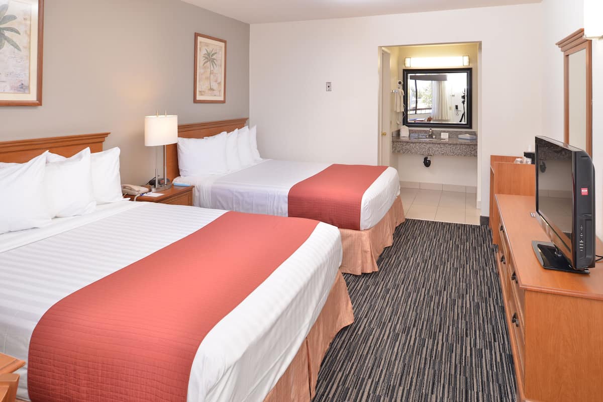 Quarto de hotel com duas camas, televisão, secretária e casa de banho visível através de uma porta aberta.