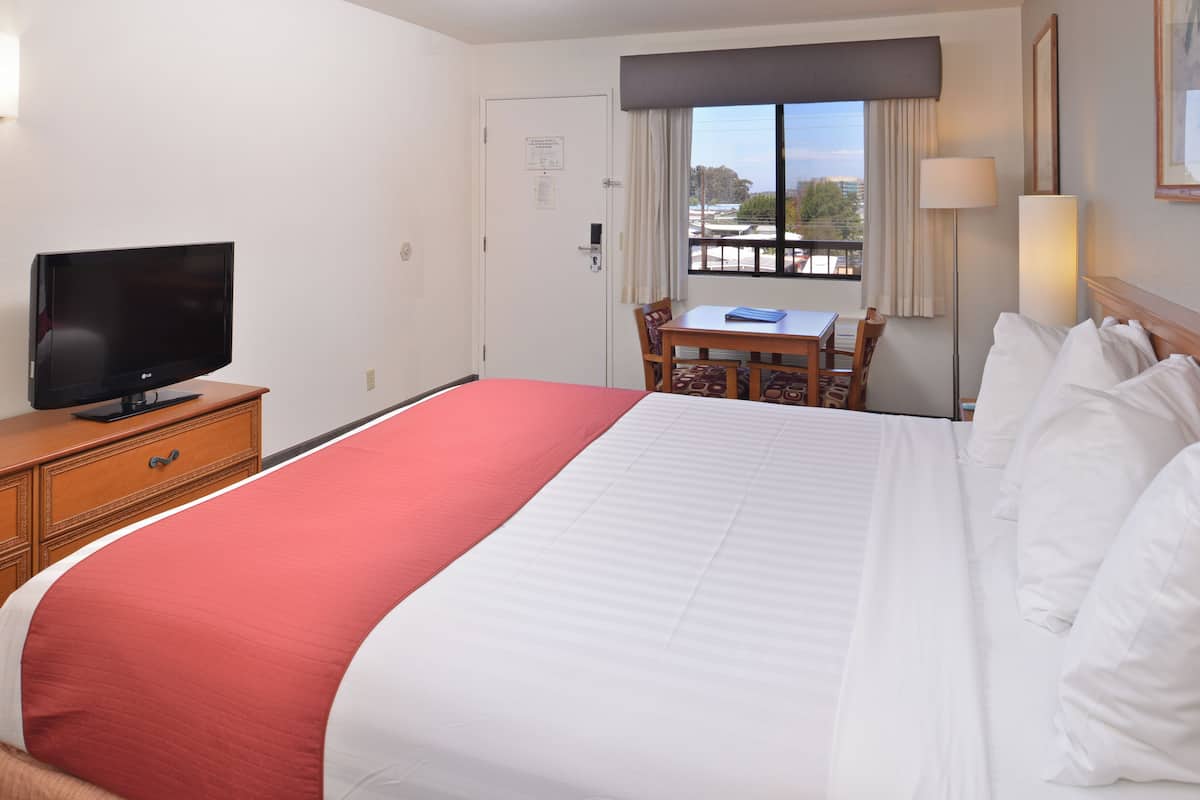 Quarto de hotel com uma cama grande, uma TV sobre uma cômoda de madeira, uma mesa de jantar com cadeiras e uma janela com vista para a cidade.