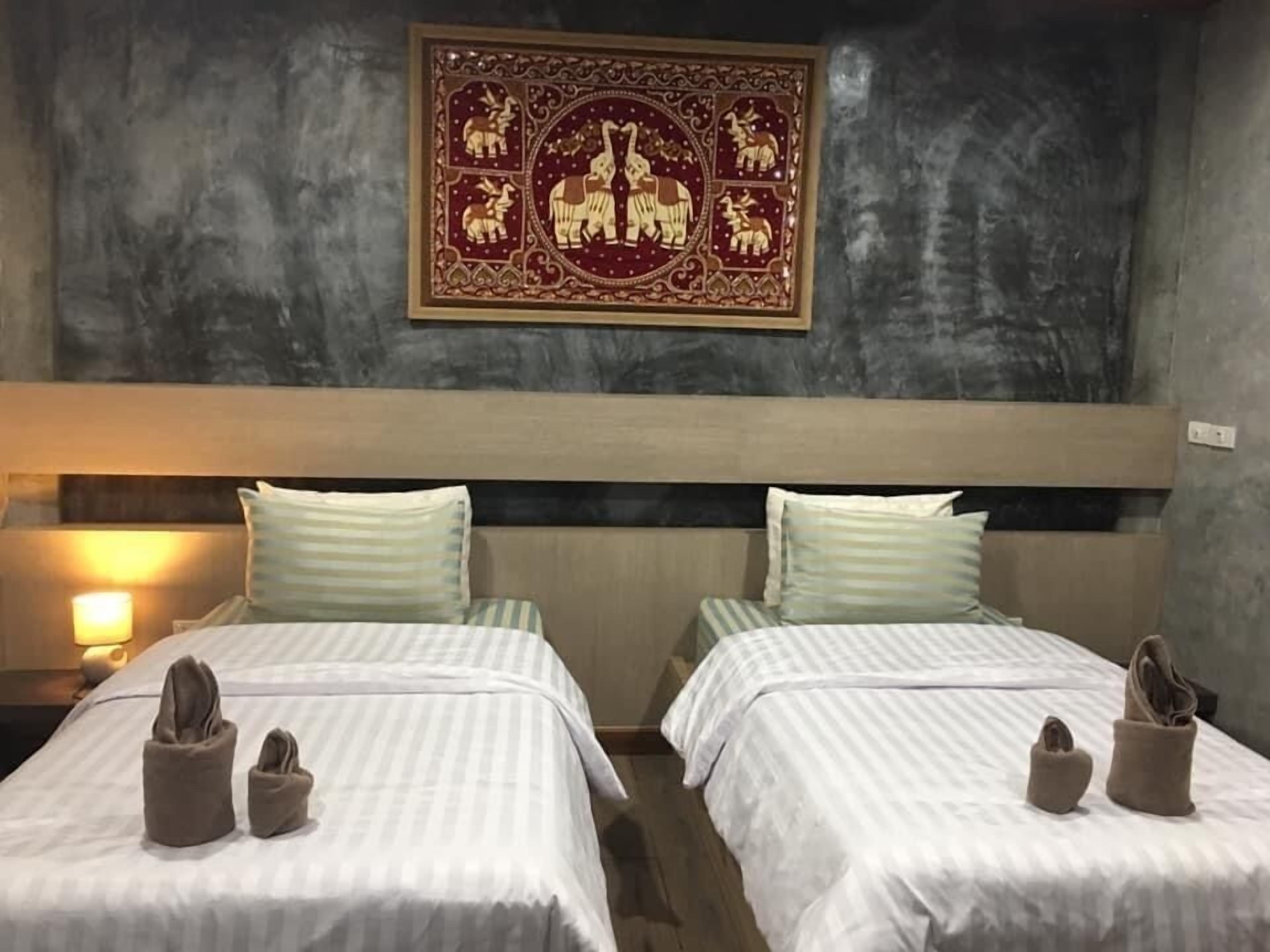 A hotel room with two beds, each with a pillow and a pair of slippers. A decorative wall hanging is above the beds.