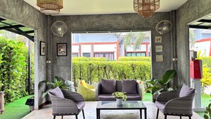 A modern interior with wicker furniture, a glass coffee table, and a view of a well-maintained garden and building outside. - Le Park 23 Boutique Resort (Roi Et)
