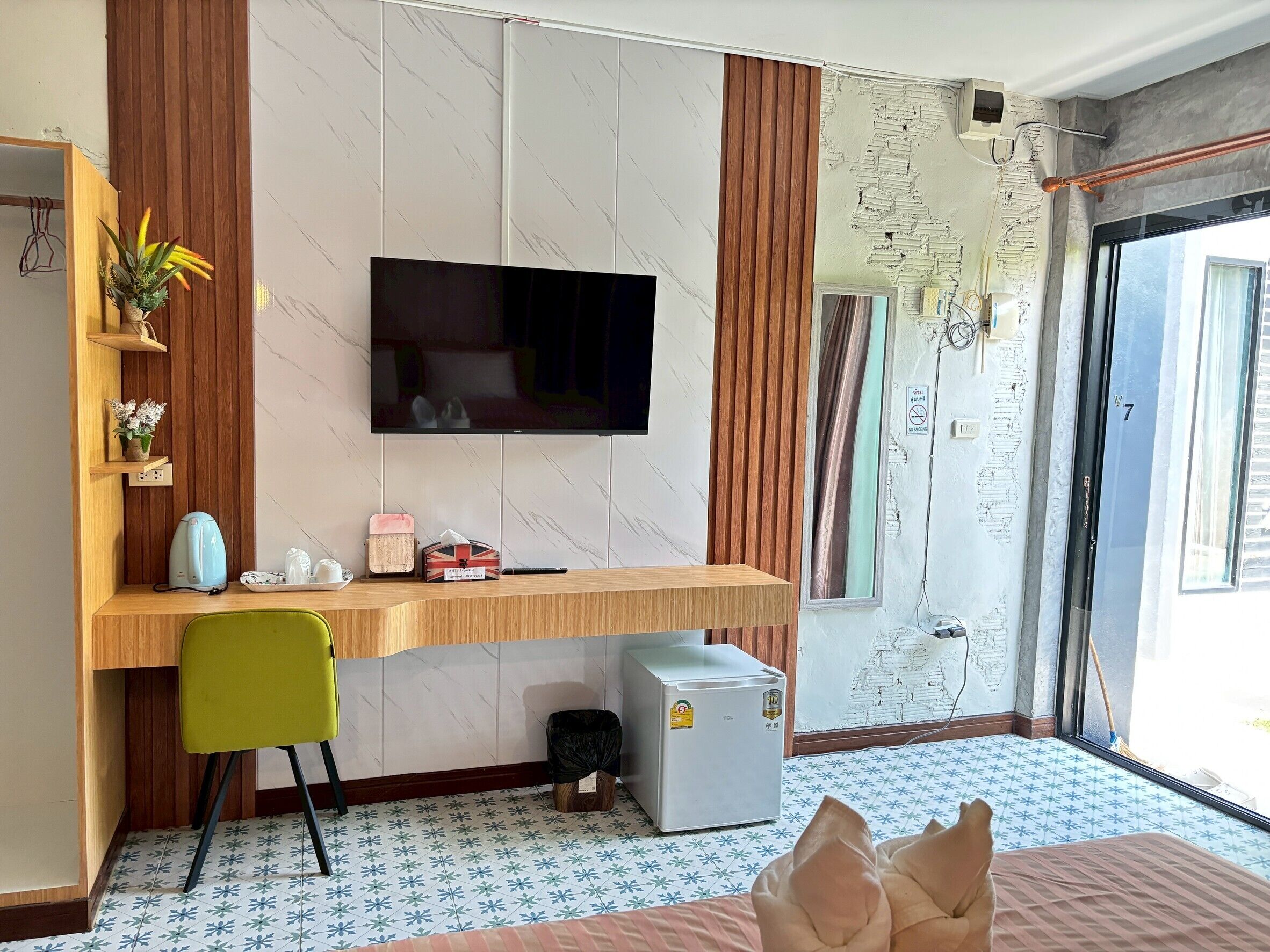 A compact room with a flat-screen TV mounted on a wall, a wooden desk, a green chair, a small refrigerator, and a window with curtains.