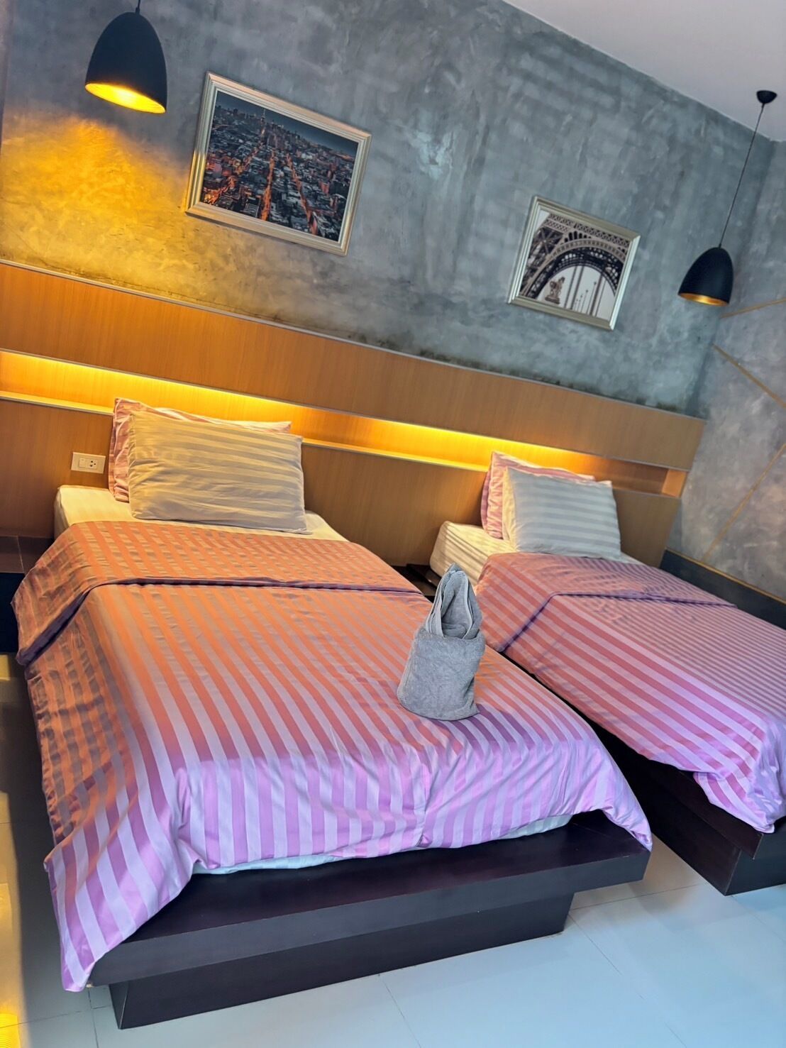 A hotel room with two beds, each with striped bedding and pillows. There are two wall-mounted lamps, a framed cityscape picture, and a framed architectural drawing on the wall.