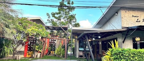 Un edificio con un espacio al aire libre techado, entrada pavimentada y áreas verdes.