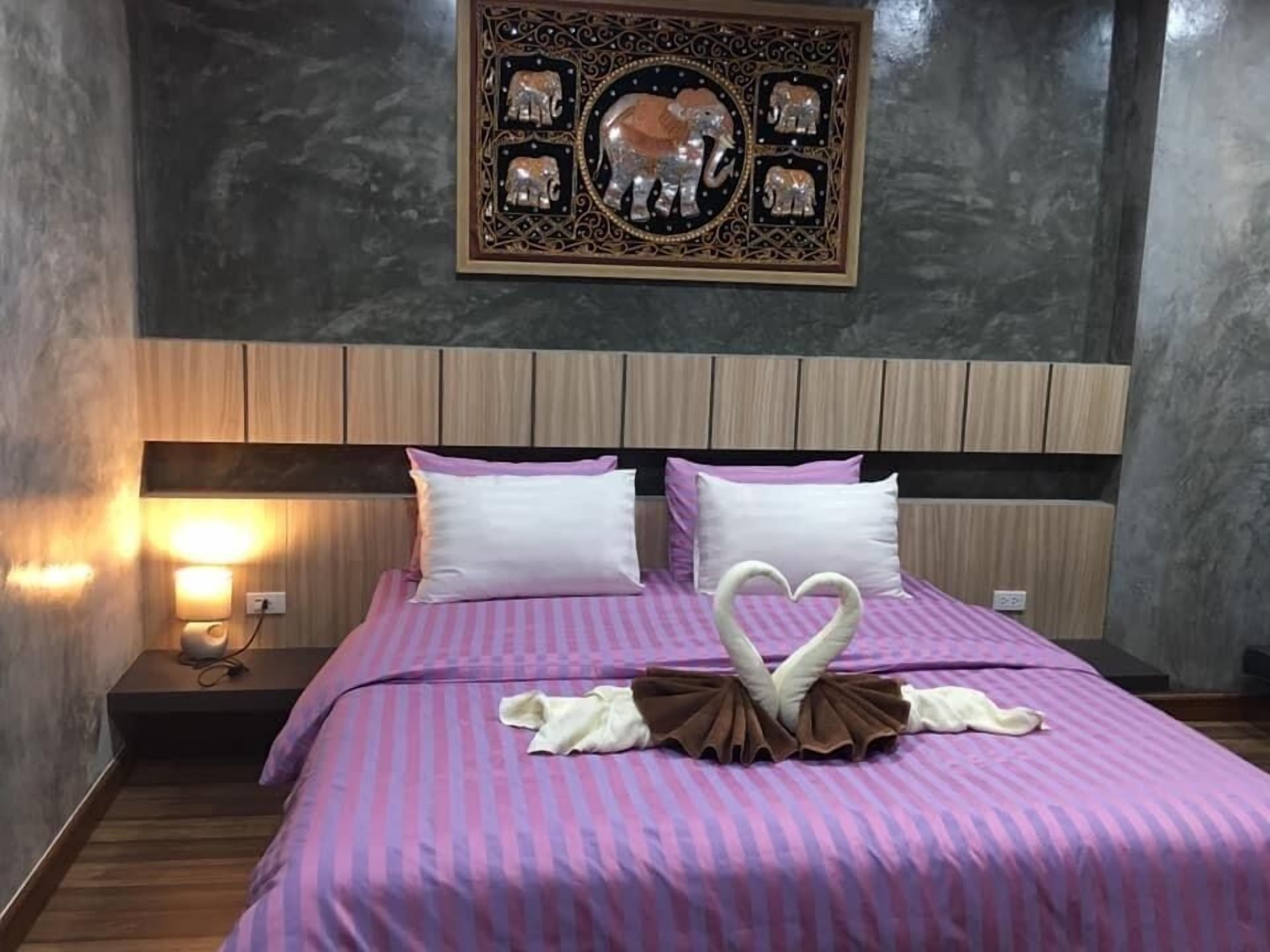 A neatly made bed with a heart-shaped towel arrangement, a wall-mounted decorative piece, and a bedside lamp.