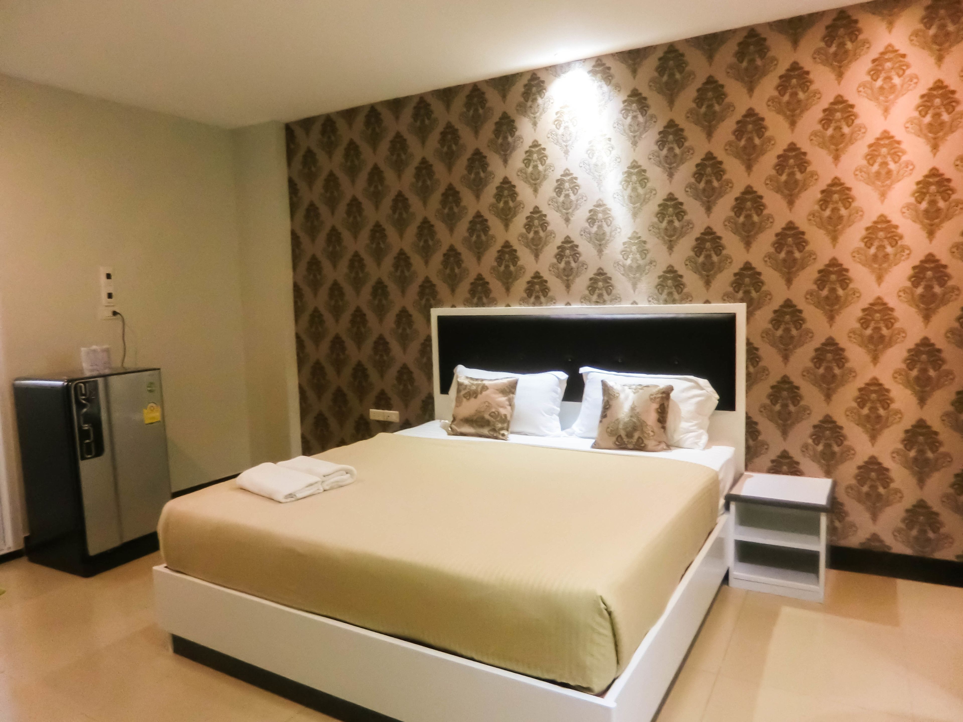 A hotel room with a bed, a nightstand, a small table, and a refrigerator.