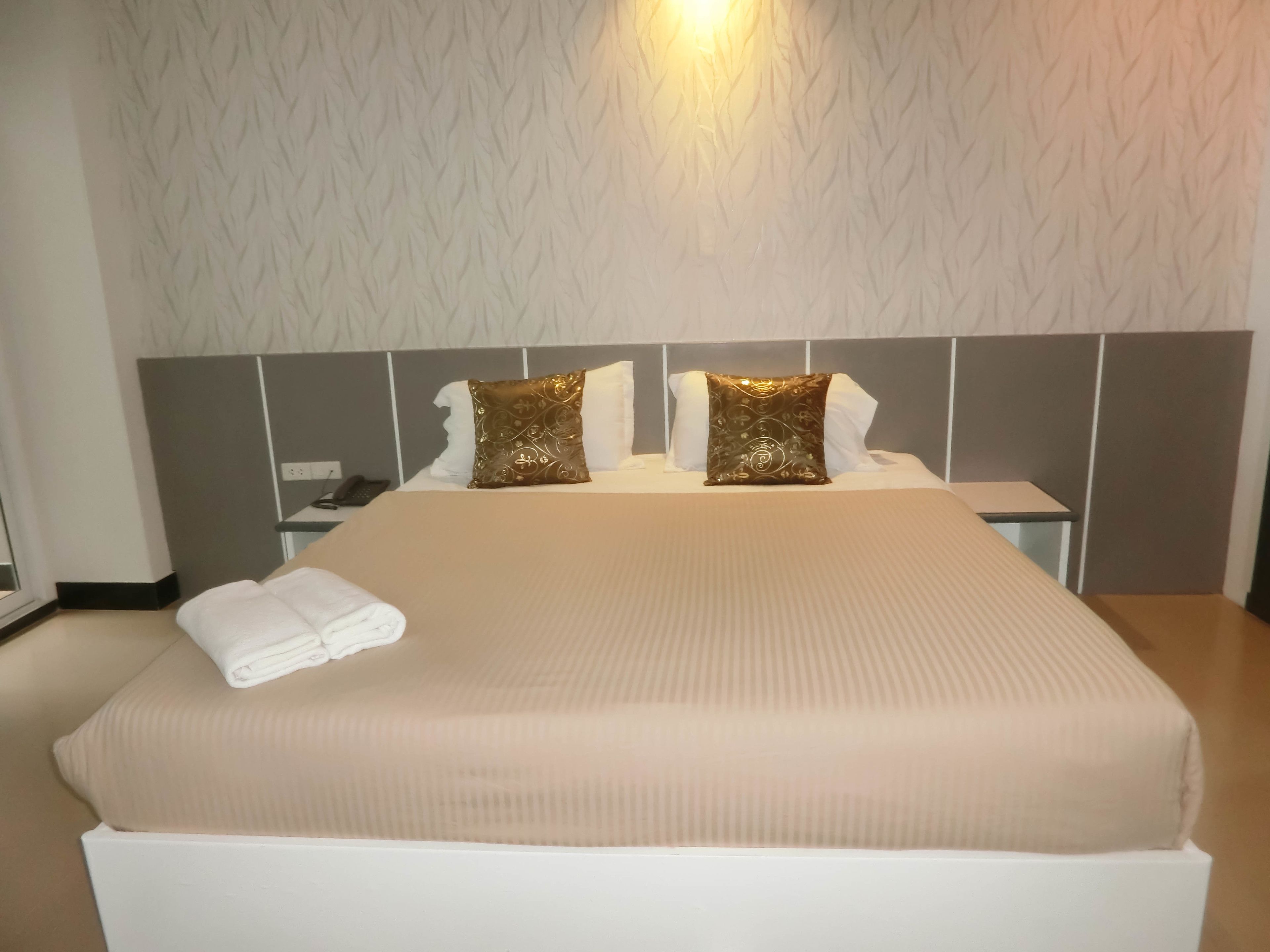 Een opgemaakt bed met een beige deken en twee decoratieve kussens, een nachtkastje met een telefoon, en een muur met een reliëf patroon.