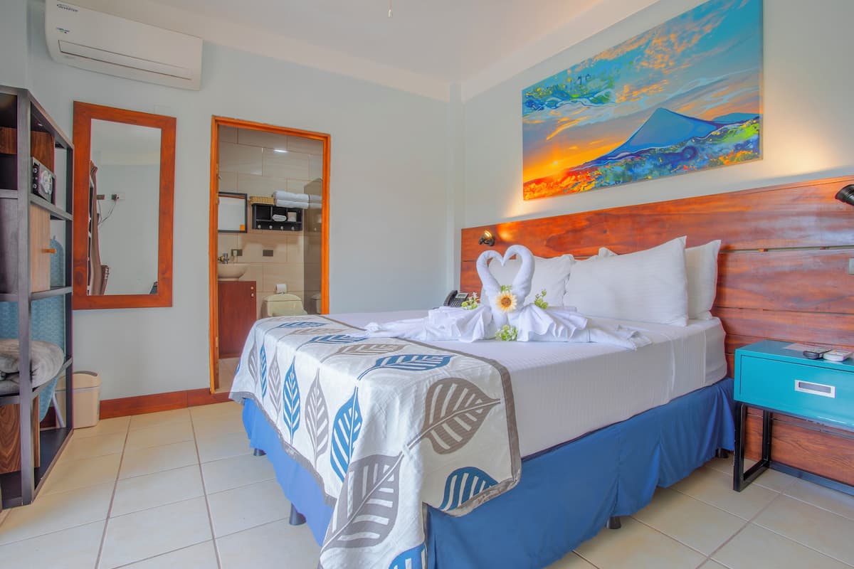 Camera d'albergo con un letto, un comodino, uno specchio, un quadro e un bagno visibile attraverso una porta aperta.