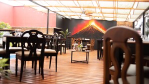 Un área de comedor al aire libre con piso de madera, mesas y sillas. Hay un mural de un volcán en la pared.