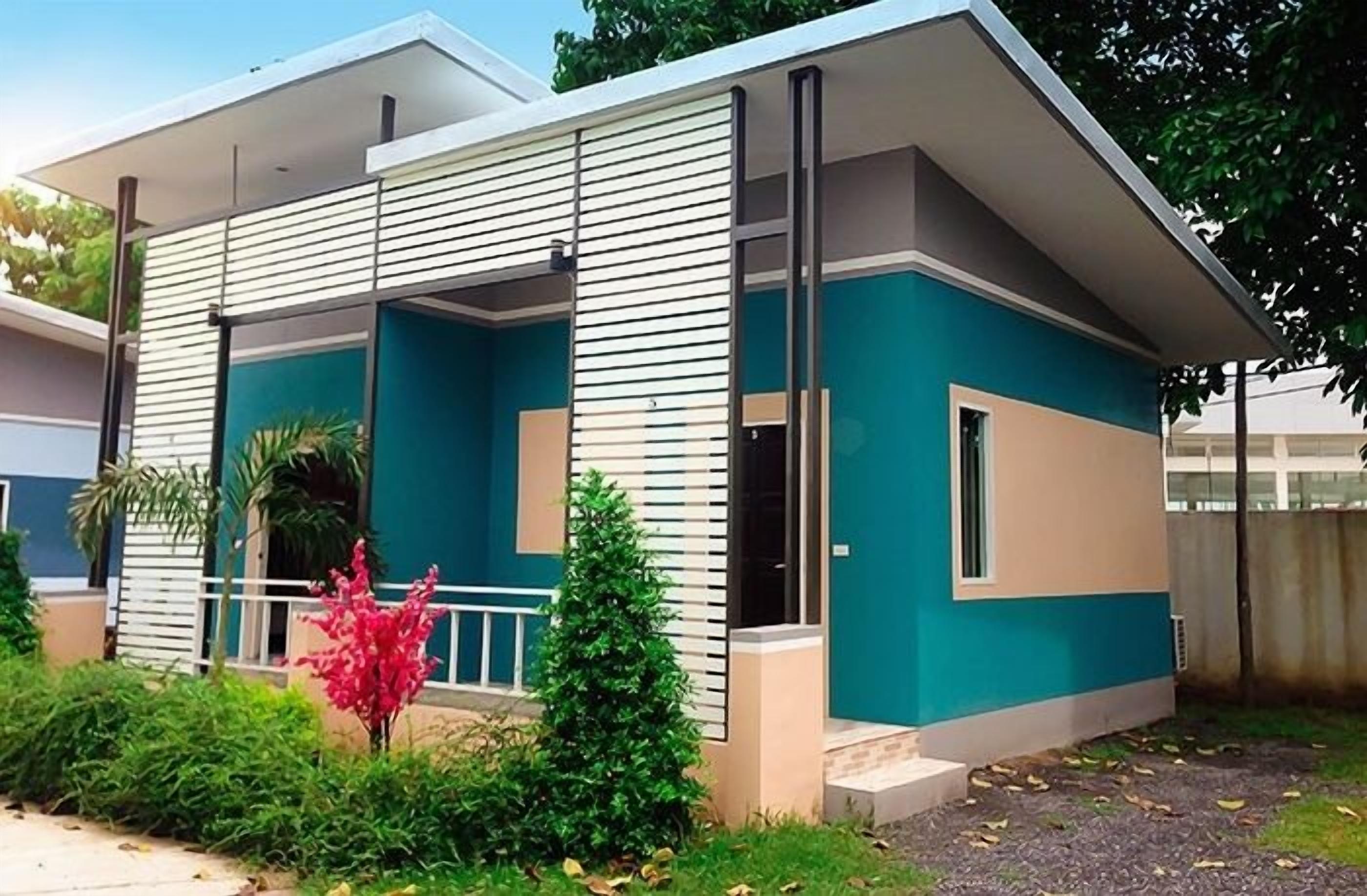 A modern two-story house with a flat roof, a balcony, and a small garden area with a red flower bush.
