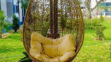 A hanging chair with a cushioned seat and back, suspended by a metal frame, in a garden setting with grass and a building in the background.