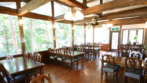 A spacious dining area with wooden tables and chairs, large windows offering a view of the surrounding forest. - PENSION & TRATTORIA Syo (Hakuba)